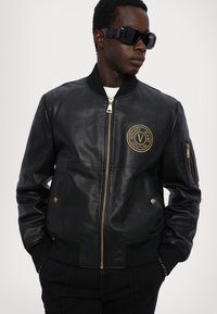 Giacca bomber in pelle nera con polsini e colletto a costine, dotata di un logo circolare dorato sul petto e tasche laterali con zip.