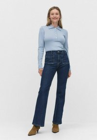 Vaaleansininen pitkähihainen polo-paita, jossa on napit, yhdistetty tummansinihin levennettyihin farkkuihin ja ruskeisiin korkokenkiin.