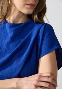 Top testurizzato blu royal con ampie maniche corte. Il tessuto appare stropicciato, riflettendo la luce. Pelle liscia visibile sul braccio.