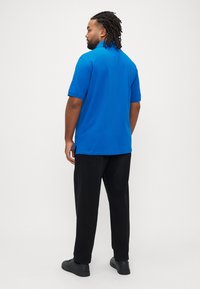 Blå polo-shirt med krave, korte ærmer og sideslidser, kombineret med sorte bukser og sorte sko, vist bagfra.