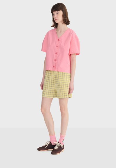 Jeune femme debout portant une chemise rose boutonnée, un short à carreaux jaunes, des chaussettes roses et des baskets marron, sur un fond uni.