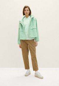 Hellgrüne Jacke mit Kapuze über einem weiß-grün gestreiften Shirt, kombiniert mit beigen Cargo-Hosen und weißen Sneakern.