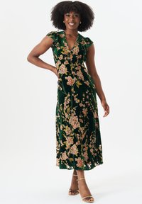 Green floral velvet dress with short ruffled sleeves, a V-neckline, and a full-length skirt featuring pink and yellow floral patterns. Gold strappy sandals.