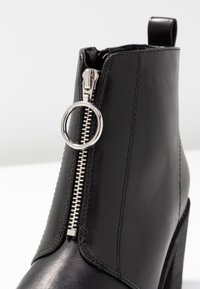 Bottine noire en cuir lisse. Dotée d'une fermeture éclair argentée avec un curseur circulaire, de détails surpiqués et d'un talon haut.