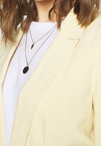 Light yellow blazer with a tailored design, featuring sharp lapels. Worn over a white t-shirt with layered silver necklaces.