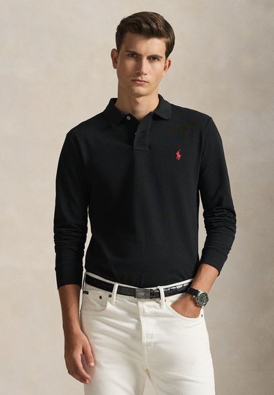 Jonge man die een zwart poloshirt met lange mouwen en rood logo draagt, witte broek, zwarte riem en polshorloge, staand met één hand in de zak.