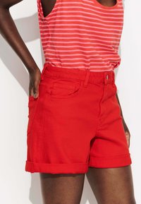 Shorts en denim rouge avec des ourlets roulés, dotés de poches avant et d'une fermeture à bouton en métal. Associés à un débardeur rayé rouge et rose.