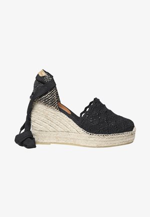 Juodas austo espadrilas su tekstiliniu viršumi, jutos pakulne ir reguliuojamais kulkšnių raiščiais. Yra raštuota dizaino ir tekstūruota baigimo.