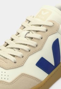 Άσπρο αθλητικό παπούτσι με μπεζ σουέντ λεπτομέρειες, μπλε λογότυπο V από καουτσούκ και ελαφρώς μπεζ κορδόνια. Διαθέτει οπές στο μπροστινό μέρος για αερισμό.