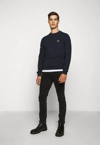 Jūras zila sporta krekls ar apaļu kaklu, teksturēts audums, neliels simbols uz krūtīm, apvienots ar melnām džinsām un melniem potīšu zābakiem.