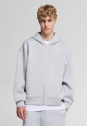 Giovane uomo con capelli biondi ricci che indossa una felpa con cappuccio grigio chiaro con zip e pantaloni della tuta abbinati su uno sfondo semplice.