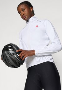 Weiße, leichte Fahrradjacke mit hohem Kragen, elastischen Bündchen und einem roten Logo. Schwarze gepolsterte Fahrradhosen und ein schwarzer Helm werden gehalten.