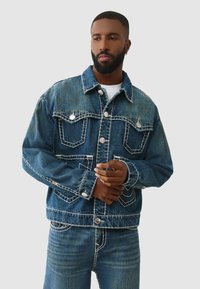 Veste en denim de couleur bleu foncé délavé, avec des surpiqûres blanches contrastantes et des boutons en métal argenté. Comprend des poches poitrine et une coupe décontractée.