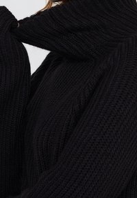 Person som drar upp den höga kragen på en tjock, svart, ribbad stickad tröja runt sitt ansikte.