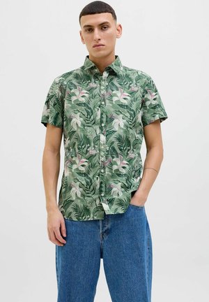 Short-sleeve green shirt featuring a tropical leaf and flower pattern, button front, and a relaxed fit, paired with blue denim pants.