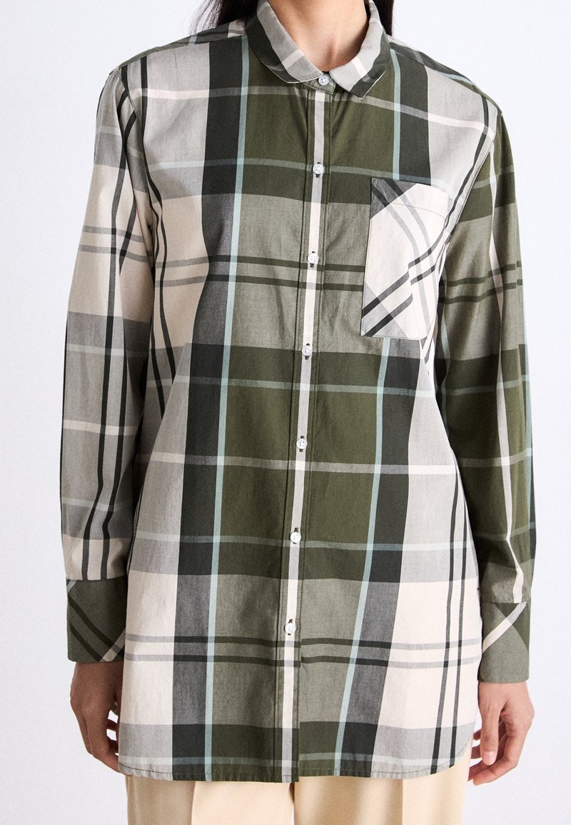 Personne portant une chemise à carreaux à manches longues boutonnée dans des tons de vert, blanc et gris avec une poche poitrine et un pantalon beige.