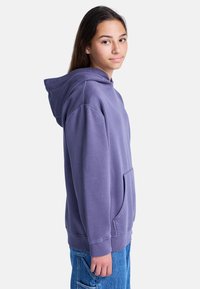 Pullover-Hoodie aus weichem, strukturiertem lila Stoff. Verfügt über eine großzügige Fronttasche und einen gerippten Saum, kombiniert mit hellblauen Jeanshosen.