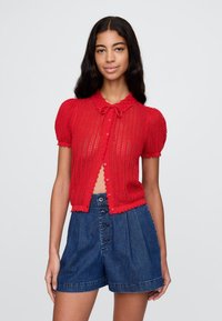 Sarkana īsām piedurknēm adīta kardigāna ar viļņotu apkakli, aizdari ar lentu un pogu priekšpusi. Komplektā ar augsta vidukļa džinsa šortiem.