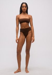 Kobieta z długimi warkoczami, ubrana w brązowe bikini bez ramiączek z majtkami z wysokim stanem, stojąca boso na jednolitym jasnym tle.