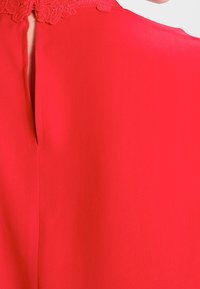 Červené oblečení s malým vertikálním rozparkem a dekorativním květinovým krajkovým lemem kolem výstřihu, ukazující texturu látky a hladký povrch.
