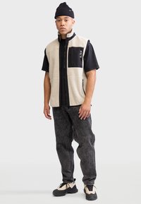 Gilet en polaire crème avec panneau noir, fermeture éclair et poche avant. Porté sur une chemise noire à manches courtes, associé à un jean foncé et des chaussures.