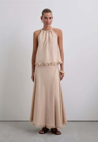 Conjunto de dos piezas en beige pálido que incluye un top halter con cuello fruncido y un dobladillo con flecos, combinado con una falda maxi que presenta un detalle de flecos similar.