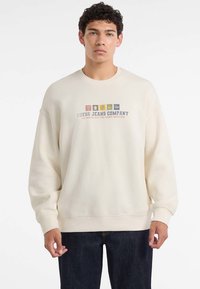 Młody mężczyzna ubrany w kremowy sweatshirt z napisem "Guess Jeans Company" oraz kolorowymi logo na klatce piersiowej, w połączeniu z ciemnymi dżinsami.