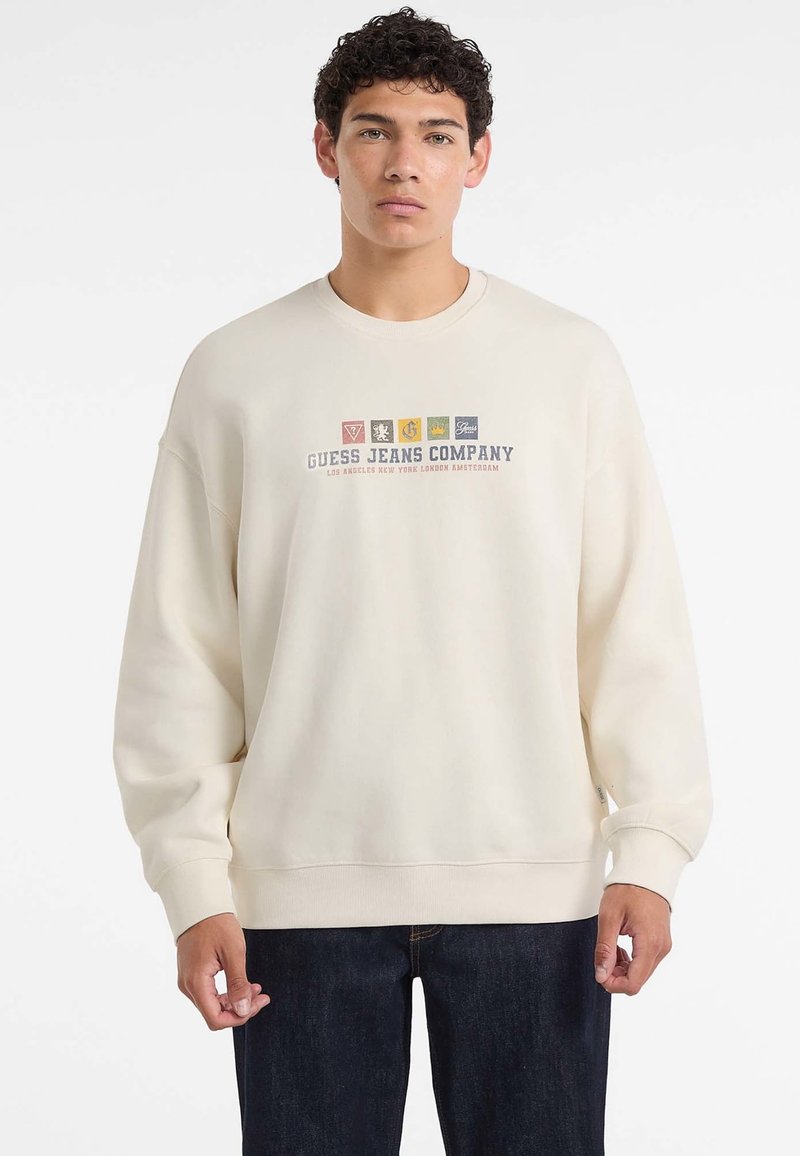 Młody mężczyzna ubrany w kremowy sweatshirt z napisem "Guess Jeans Company" oraz kolorowymi logo na klatce piersiowej, w połączeniu z ciemnymi dżinsami.