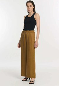 Zwart tanktop gecombineerd met losse, hoge bruine broeken van zacht materiaal. Schoeisel bestaat uit zwarte sandalen met bandjes.