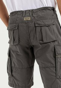 Graue Cargo-Shorts mit mehreren Taschen, elastischem Taillenbund und einem Etikett mit der Aufschrift "FÜR TÄGLICHE ABENTEUER SEIT 1977 GEMACHT." Leichter Stoff.
