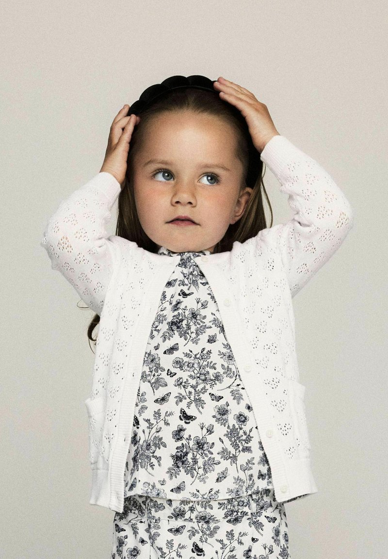 Vit, öppet stickad cardigan med blommönstrad skjorta under. Barnet håller i ett svart hårband, har långt hår och blå ögon. Enkel bakgrund.