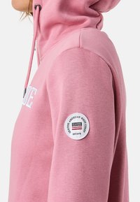 Rosa Kapuzenpullover mit Kängurutasche, Zugbandverschluss und einem runden Aufnäher mit der Aufschrift "Seaside American West Coast" am Ärmel.