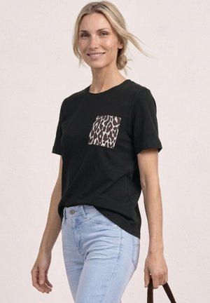 Lächelnde Frau mit zurückgebundenen blonden Haaren trägt ein schwarzes T-Shirt mit Leopardenmuster-Tasche und hellblaue Jeans und hält den Griff einer braunen Tasche.