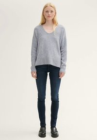 Vaaleanharmaa v-kaula-aukollinen neule, jossa on ribbineuloshelma, yhdistetty tummaan siniseen kapealahkeisiin farkkuihin. Mallilla on jalassaan mustat korolliset kengät.