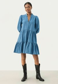 Denim suknelė su sluoksniuota apatine dalimi, V formos iškirpte ir ilgomis pūstomis rankovėmis. Mėlyna spalva su lygiu paviršiumi. Derinama su juodais ilgaauliais.