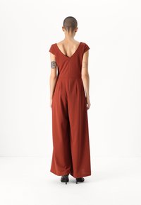 Jumpsuit boje rđe s kratkim kapicama, dubokim V izrezom na leđima i širokim nogavicama. Tekstuirani materijal s skrivenim patentnim zatvaračem na leđima.