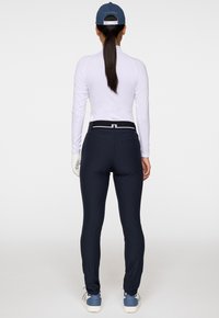 Kvinna med lång hästsvans iklädd en vit långärmad topp, marinblå byxor, blå keps, vita handskar och blå sneakers som står vänd bortåt mot en vit bakgrund.