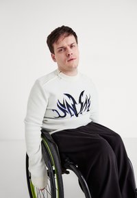 Λευκό πλεχτό πουλόβερ με αφηρημένο σχέδιο σε μαύρο και μπλε, με φερμουάρ στους ώμους. Μαύρο παντελόνι, άτομο σε αναπηρικό αμαξίδιο. Ουδέτερο φόντο.