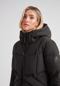 Ung kvinde med blondt hår iført en sort polstret vinterjakke med hætte, lynlås og et logo-patch på ærmet.