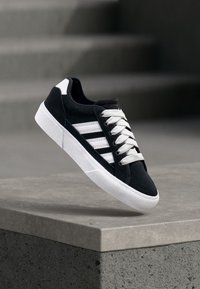 Black suede sneaker with white laces and three white stripes on the side. Features a white rubber sole and rounded toe design.