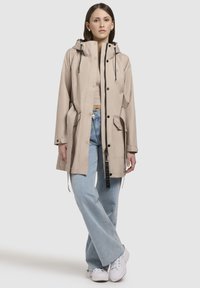 Beige waterdichte jas met een capuchon, voorknopsluiting en aantrekkoorden; gecombineerd met lichtblauwe wijde jeans en witte sneakers.