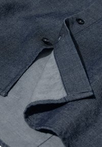 Marineblå denimstof med en glat, struktureret overflade. Har en knaplukning og en kontrasterende lys grå indvendig foring. Foldet i en vinkel.