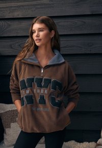 Brun fleece-trøje med grå krave, med et stort "NYC"-motiv, blød tekstur og lynlås i halsen. Sorte leggings fuldender outfittet.