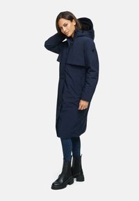 Manteau long bleu marine avec capuche, à l'avant doublé, poches latérales et panneau texturé dans le dos. Porté avec un jean et des bottes noires.