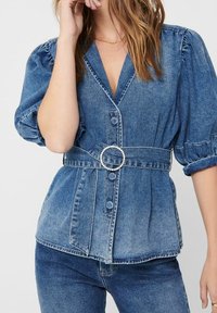 Denim top with a V-neck, puffed sleeves, and buttons, featuring a cinched waist with a round, jeweled belt. Blue color with textured fabric.