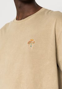 Béžové tričko s malými barevnými vyšitými houbami na hrudi, nošené osobou s tmavou pletí.