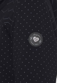 Sudadera de color navy con pequeños lunares en azul claro. Presenta un parche texturizado negro con un logotipo en relieve y un detalle de costura decorativa.