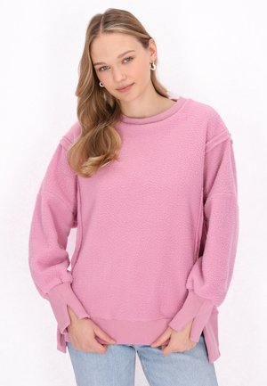 Felpa rosa pelosa con collo e polsini a coste, spacco laterale e spalle scese. Abbina con jeans azzurri chiari.
