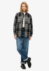 Sorte og hvide ternet skjorte med to brystlommer, lavet af en bomuldsblanding; båret over en hvid top, parret med opsmøgede blå jeans og sorte støvler.