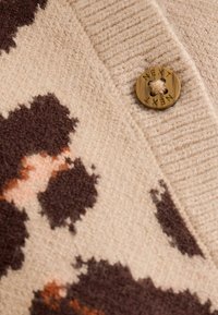 Pulover din țesătură tricotată, având un fundal bej cu modele abstracte maronii și portocalii deschise, finisat cu un nasture rotund maroniu pe care scrie "NEXT."